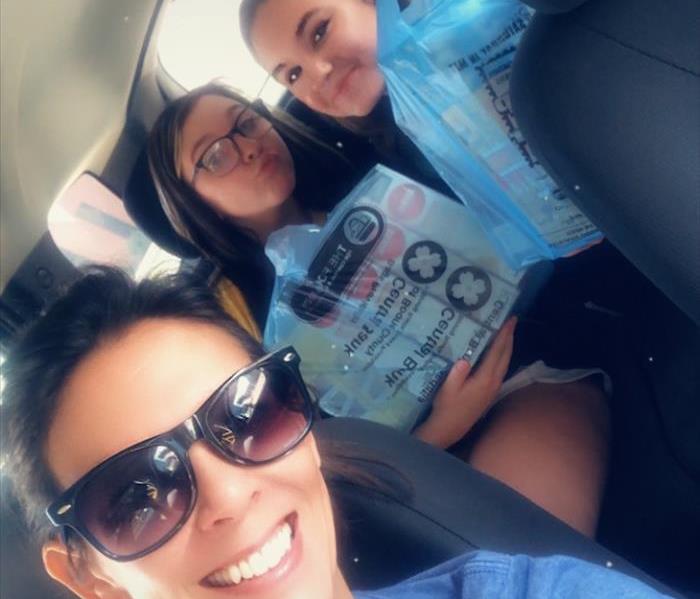 Three women in a car, two holding up blue grocery bags filled with food